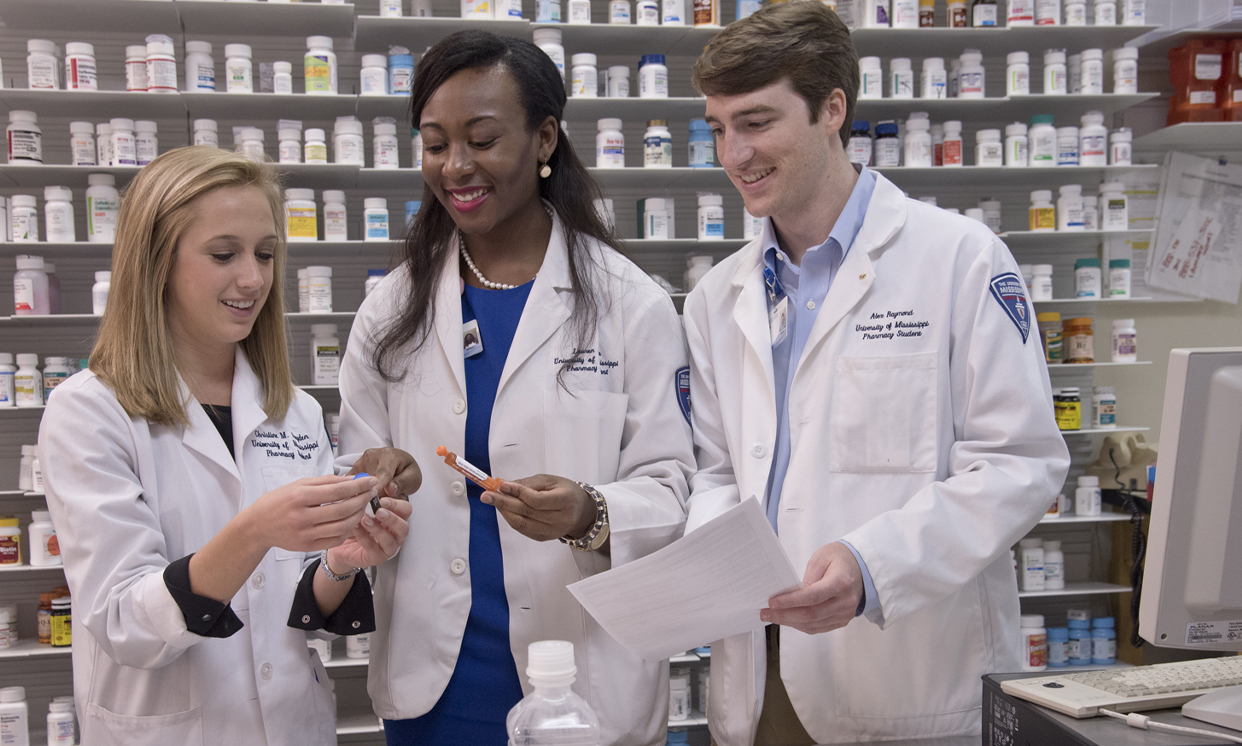 Three pharmacy students standing in front of a wall of pill bottles